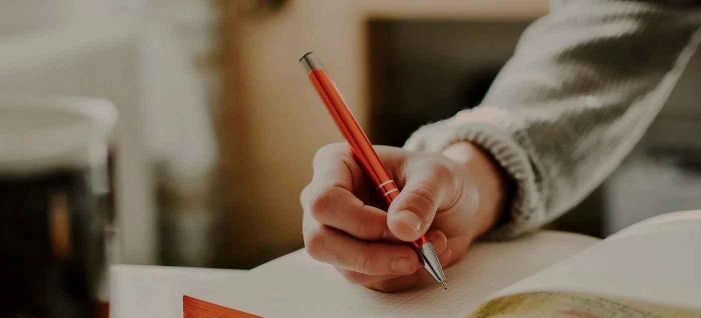 gros plan sur une main écrivant au stylo orange dans un carnet, symbolisant la correction méthodique des erreurs d'un site web